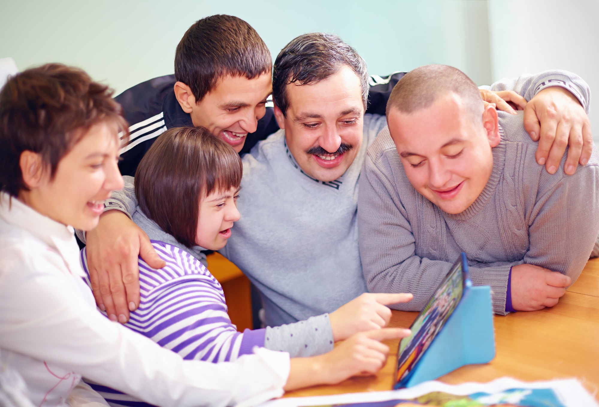 A group of five people, including a girl with Down syndrome, joyfully interact while looking at a tablet on a table, showcasing shared learning and connection.